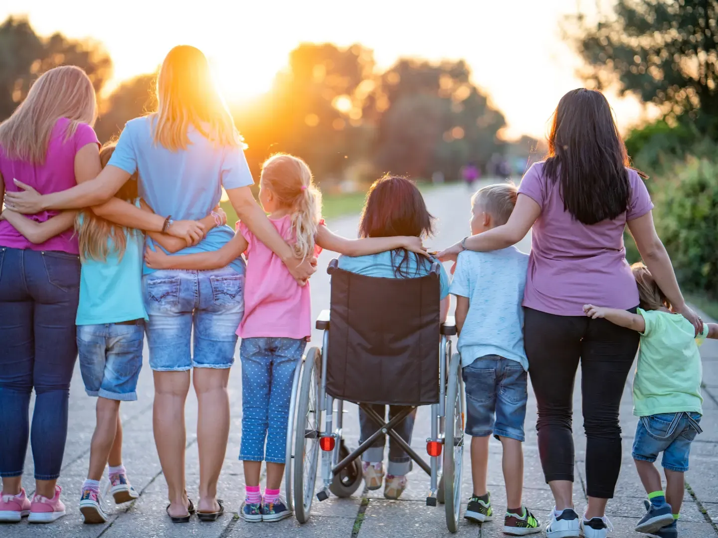 Erwachsene und Kinder stehen in einer Reihe und umarmen sich. In der Mitte befindet sich ein Kind im Rollstuhl.