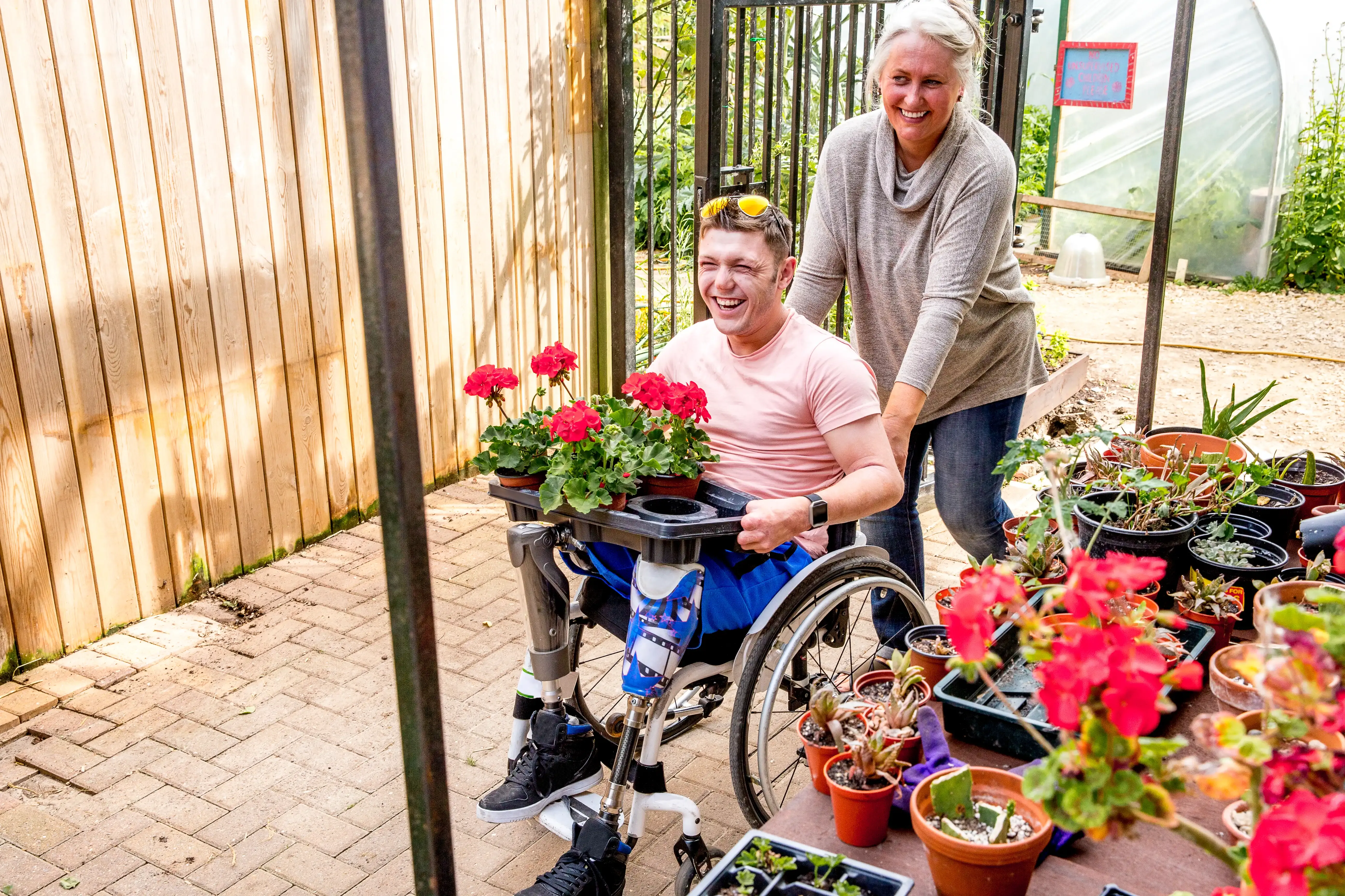 Ein Mann im Rollstuhl hält Blumensetzlinge in der Hand. Dahinter steht seine Assistentin, die ihn unterstützt.