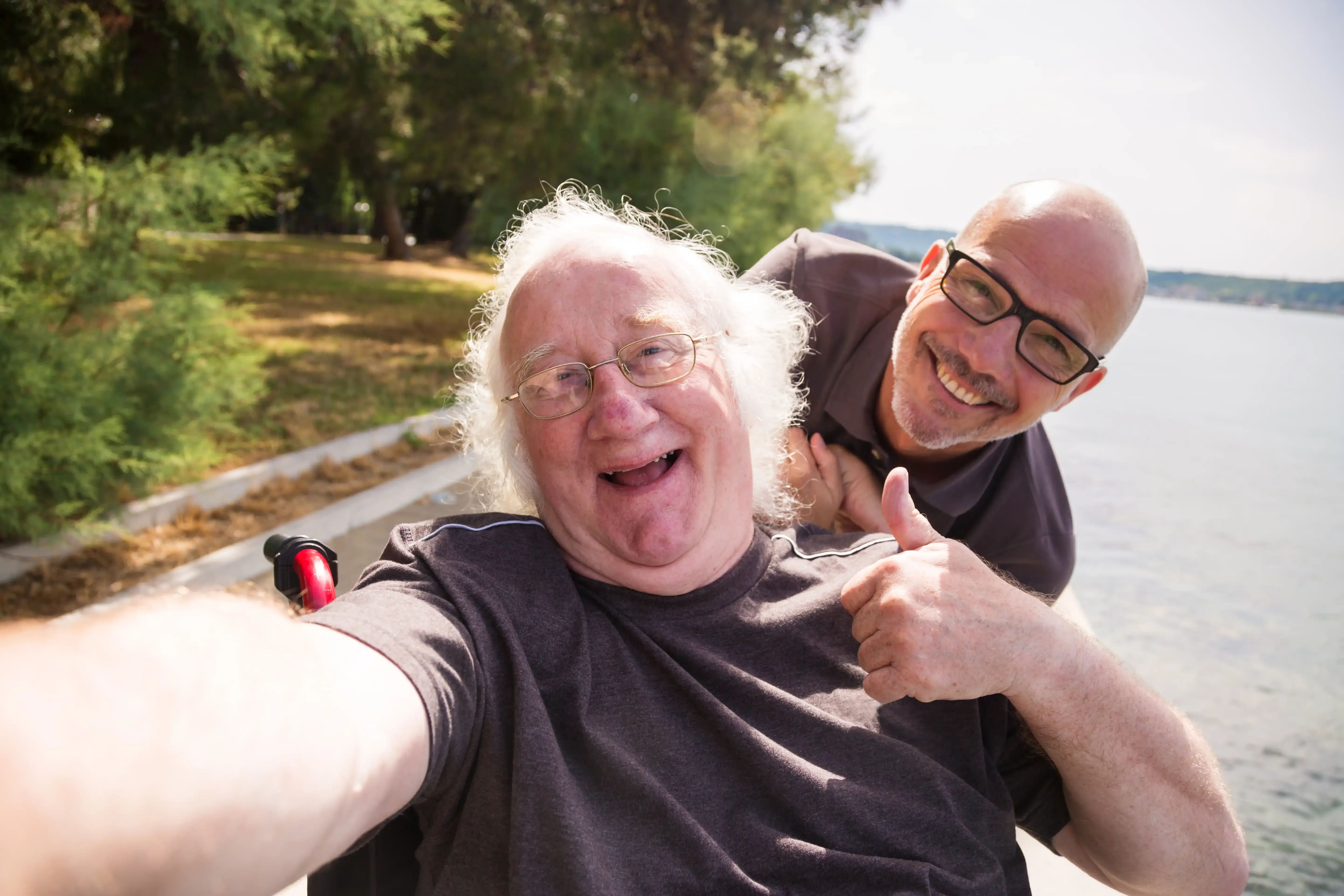 Zwei Personen machen ein Selfie am Ufer eines Sees, eine Person sitz im Rollstuhl und zeigt Daumen hoch, im Hintergrund Bäume und Wasser.