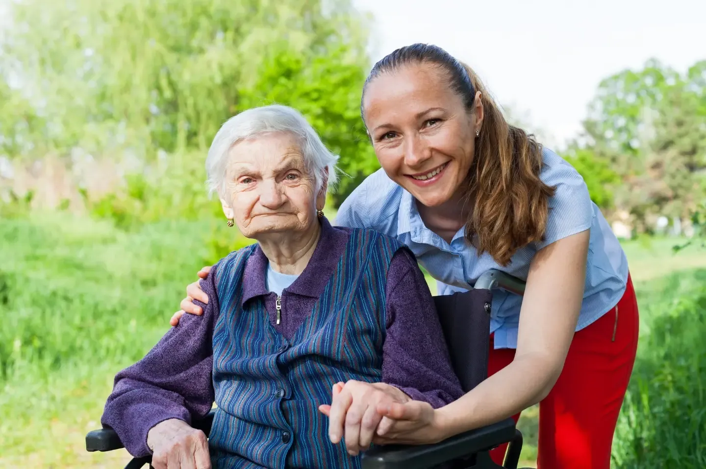 Ältere Person im Rollstuhl hält die Hand einer jüngeren Frau, die sich zu ihr hinunterbeugt im Garten