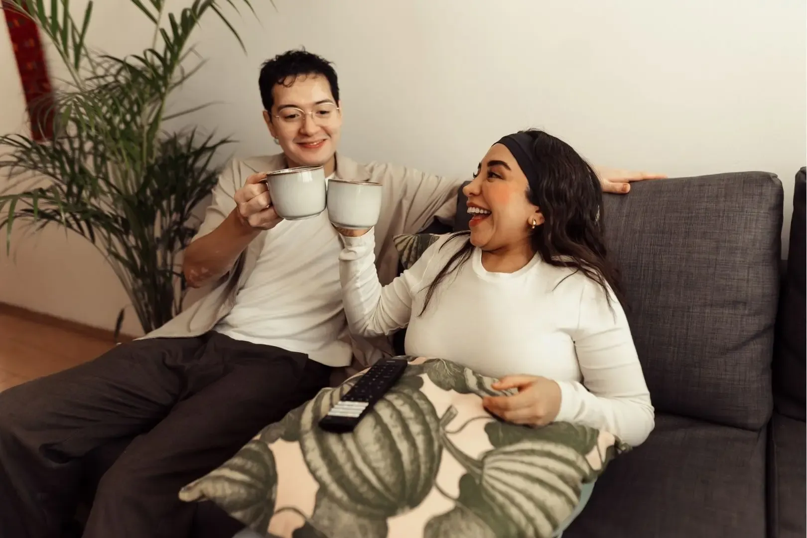 Zwei Personen sitzen auf einem grauen Sofa, halten weiße Tassen und stoßen an.